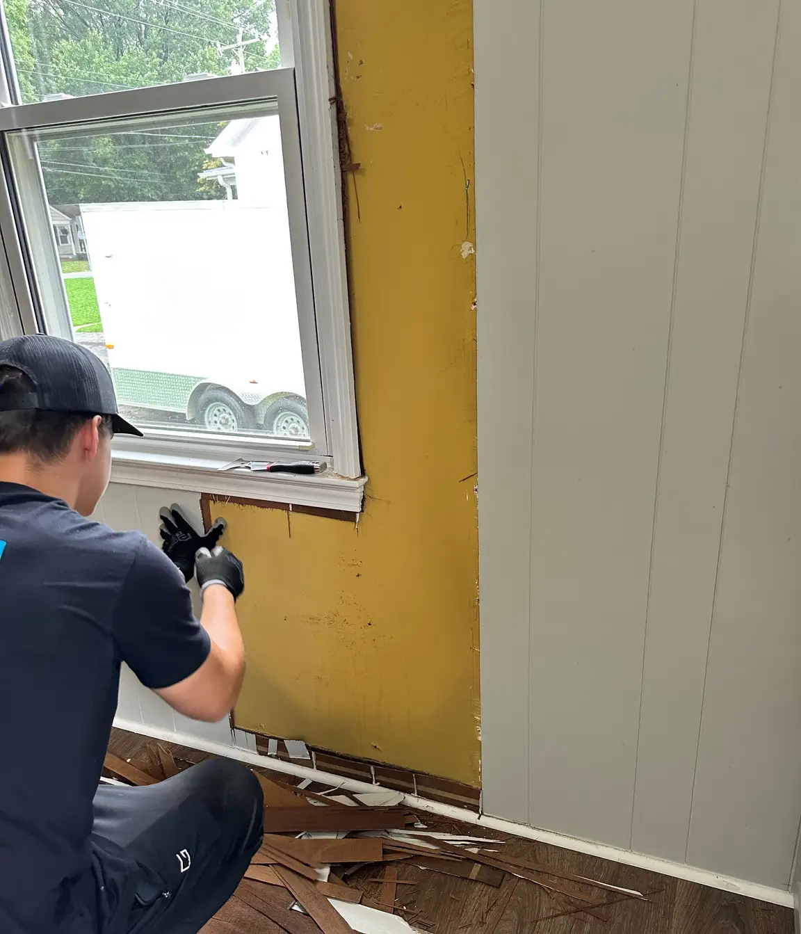 Contractor removing painted wood paneling around a window, revealing the older wall surface and hidden conditions behind it.