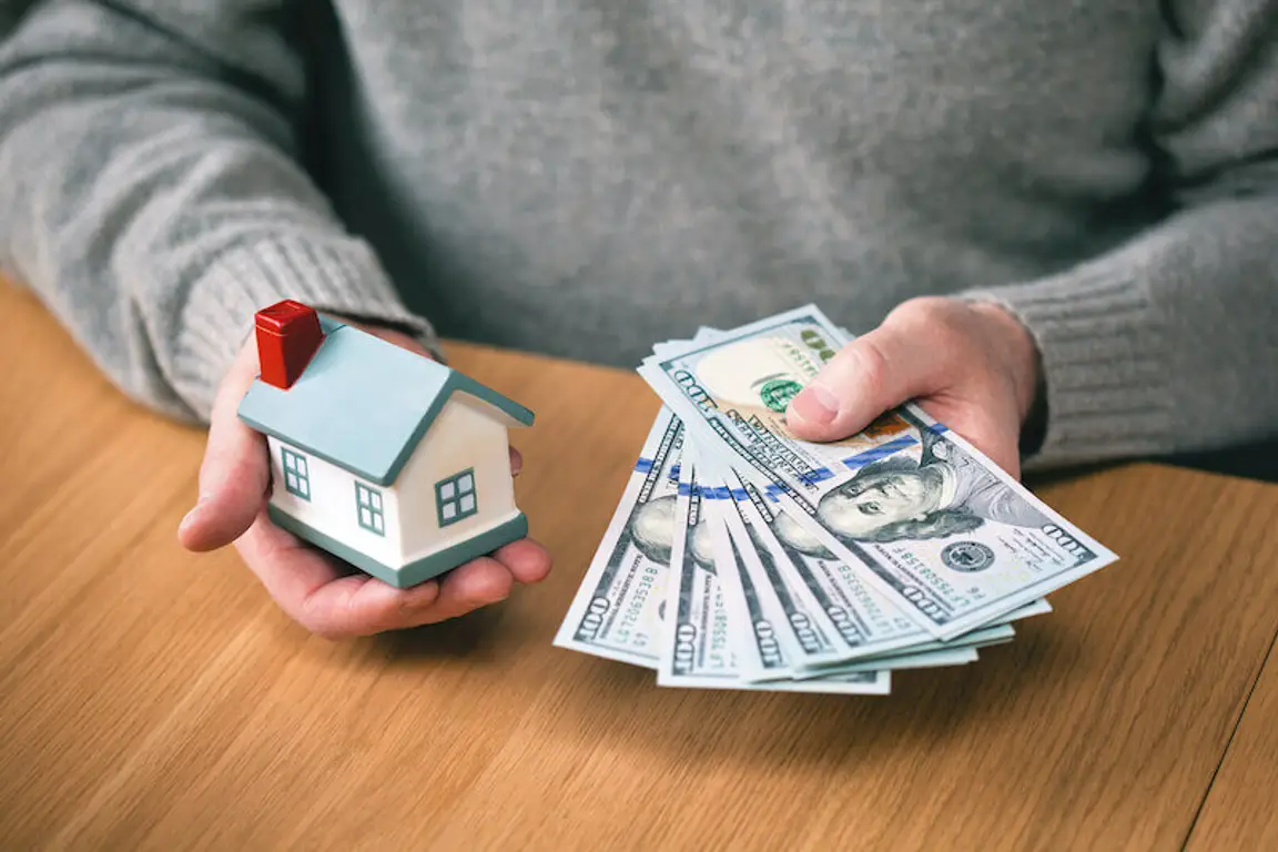 hands holding new hundred-dollar bills and toy house