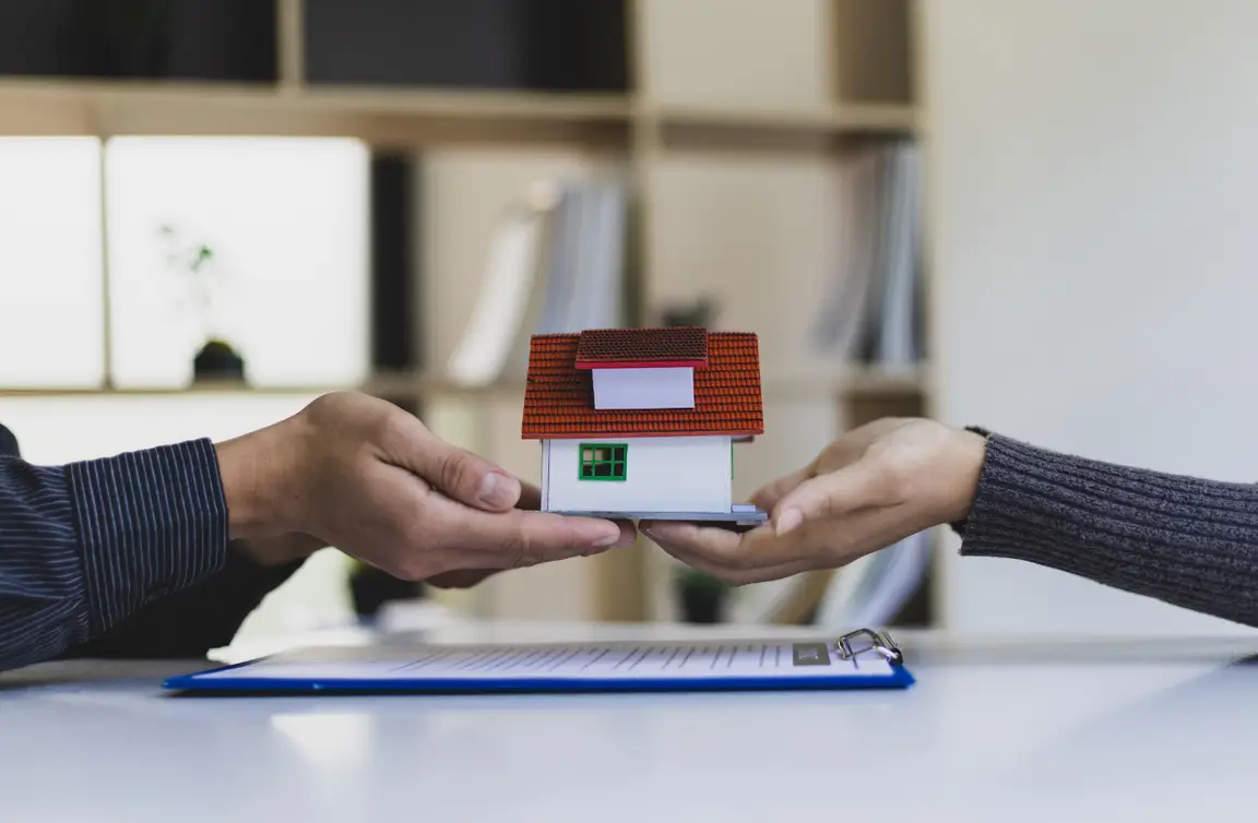 Stock photo for inherited house: two pairs of hands holding a small model house over title transfer paperwork.