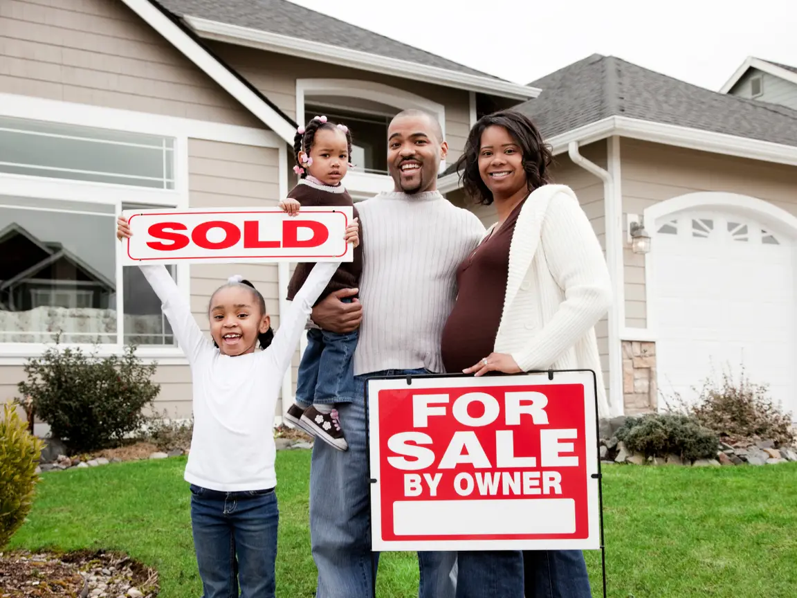 A husband, wife, and two daughters in the front yard of their house. They stand behind a "For Sale By Owner" sign, and one of the daughters holds a "Sold" sign.