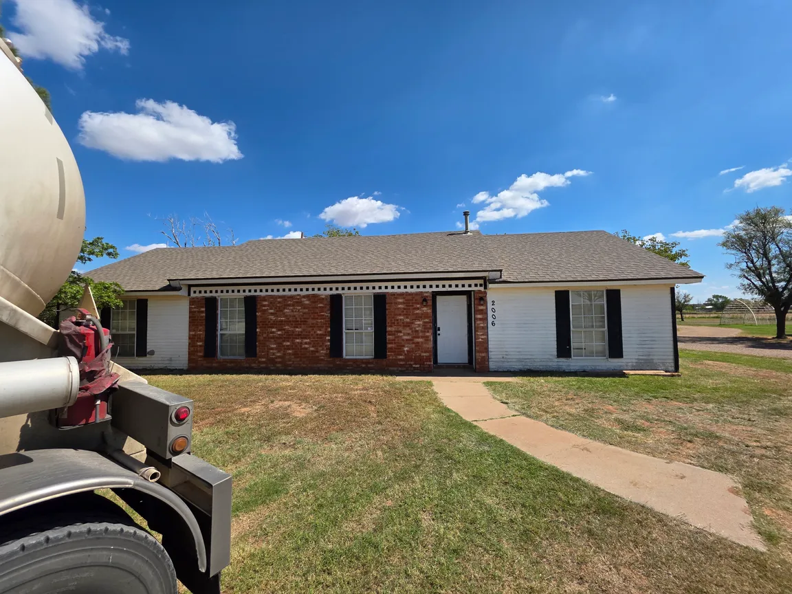 A single-story brick and white-siding house with black shutters under a bright blue sky in Lubbock, Texas. A curved concrete walkway leads to the front door, and a large utility truck is partially visible on the left side of the frame. The yard is dry with patches of grass, and open land with trees sits in the background.