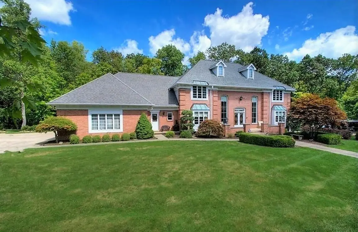 A photograph of a custom-built luxury home within Connecticut Woods subdivision, located along the cul-de-sac street of Connecticut Woods Drive in Hudson, Ohio 44236. This 2-story home features a stately red full brick façade with copper roofed bay windows, a double entry front door, a courtyard surrounded by a brick pony wall, lush landscaping, and a wooded backyard. The custom-built homes of Connecticut Woods were constructed between 1986-1999 and range from 4,000-6,800 square feet with 3-5 bedrooms and 3-5.5+ bathrooms.