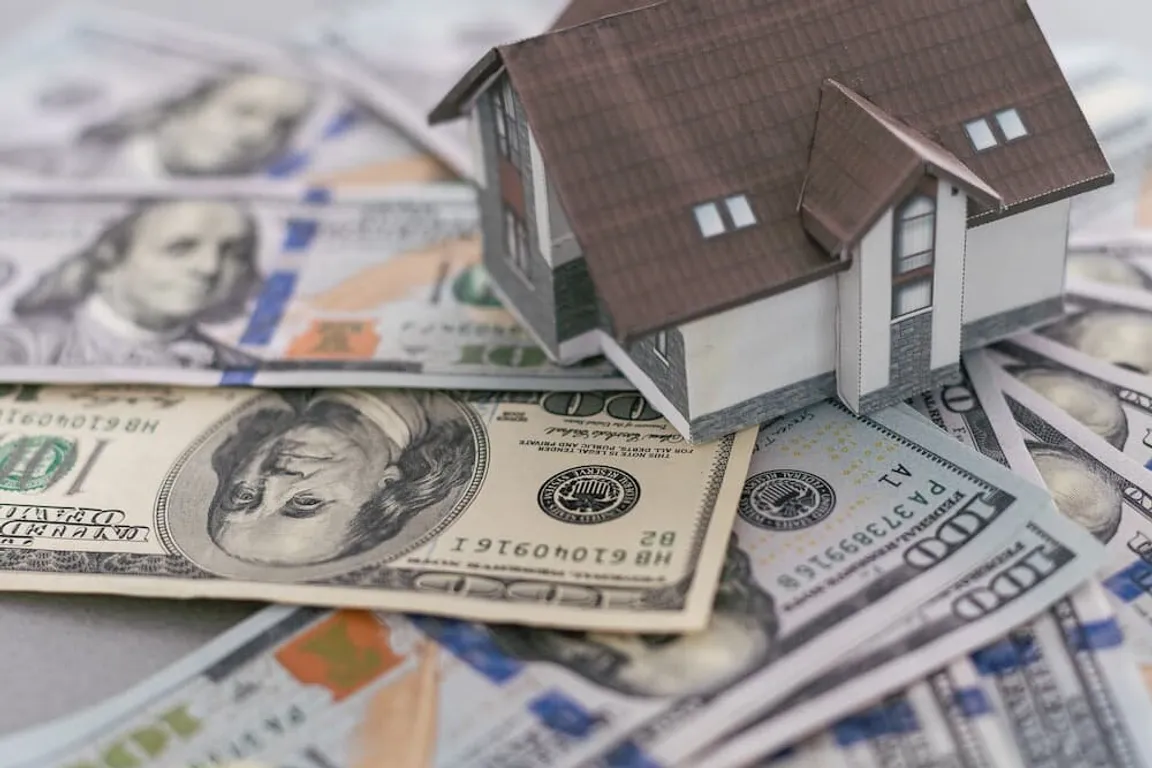 Miniature model house resting on a scattered pile of US hundred dollar bills, illustrating the financial transaction and ease of selling a house directly for cash for houses with minimal homeowner maintenance.