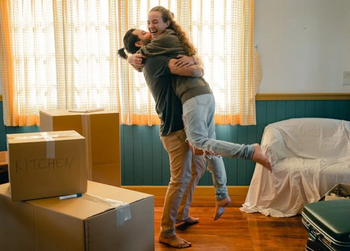 Two people embrace joyfully in a room with moving boxes, indicating they are moving out before selling their home
