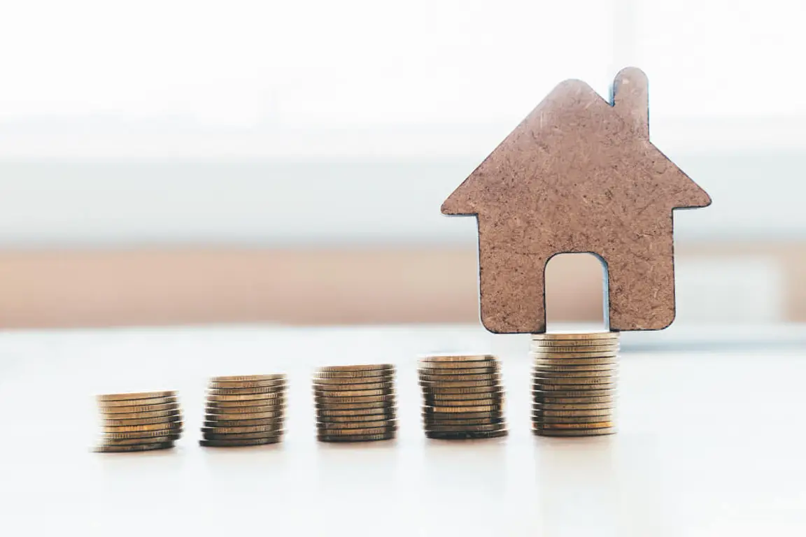 Stack of coins with a house made of cardboard in the background.