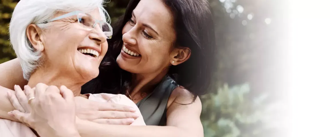 An older woman is embraced from behind by her daughter. They smile at one another.