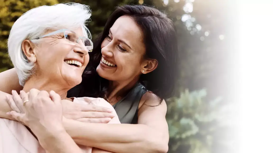 An older woman is embraced from behind by her daughter. They smile at one another.
