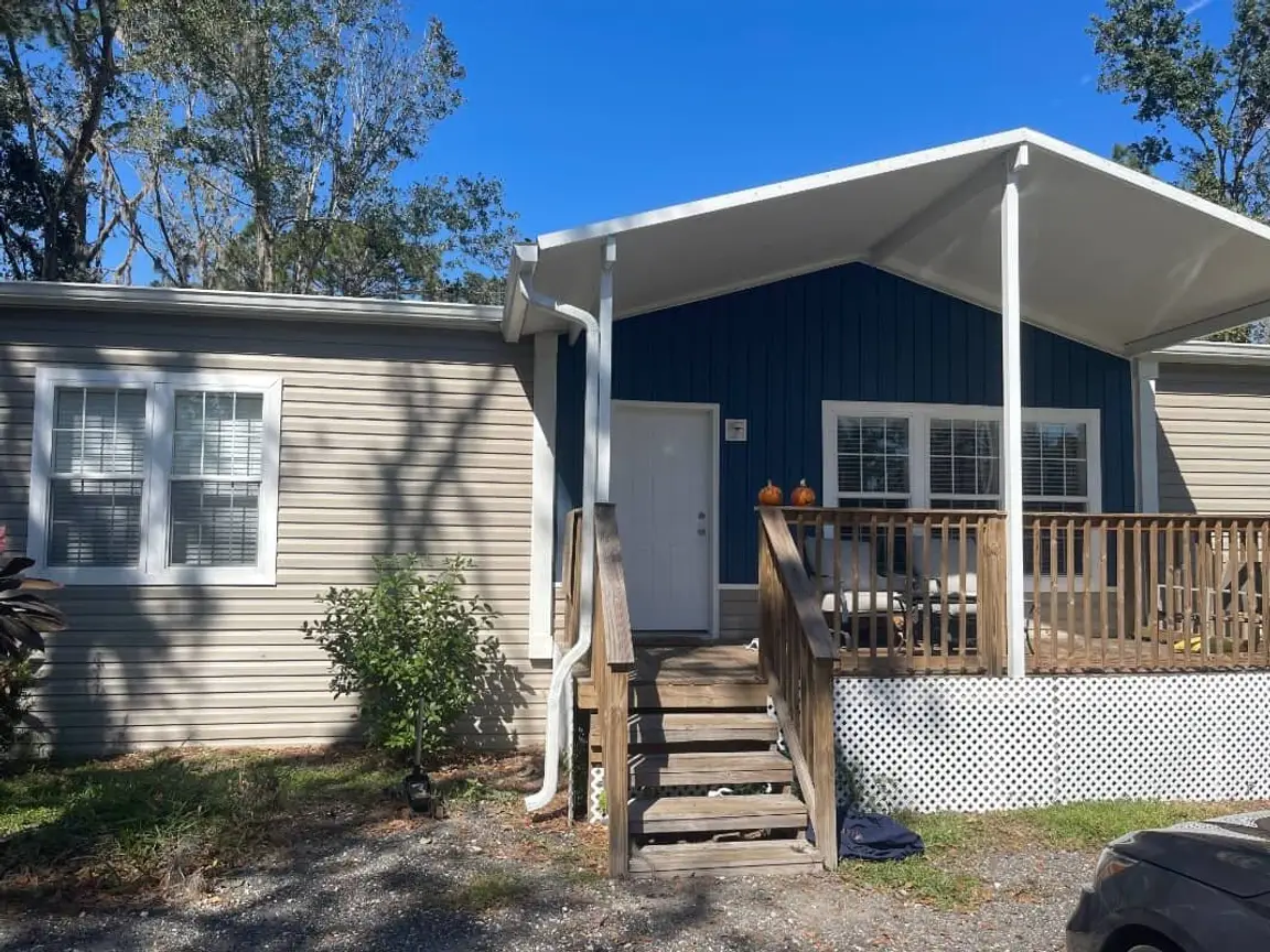 Well kept Florida mobile home with beige siding, a navy gable end and a covered wooden porch with white lattice skirting