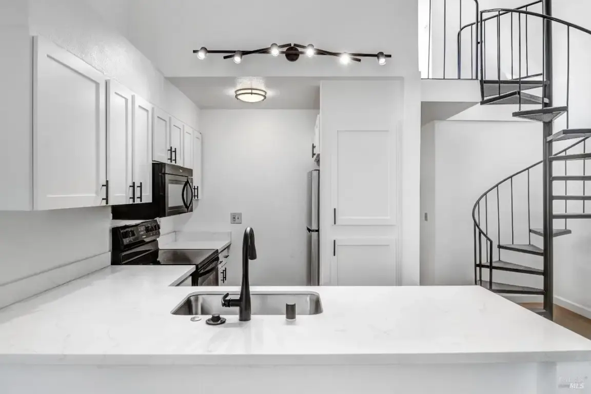Interior of a Windsor, CA condo kitchen with white cabinets, quartz counters, black fixtures, stainless appliances, and a metal spiral staircase.