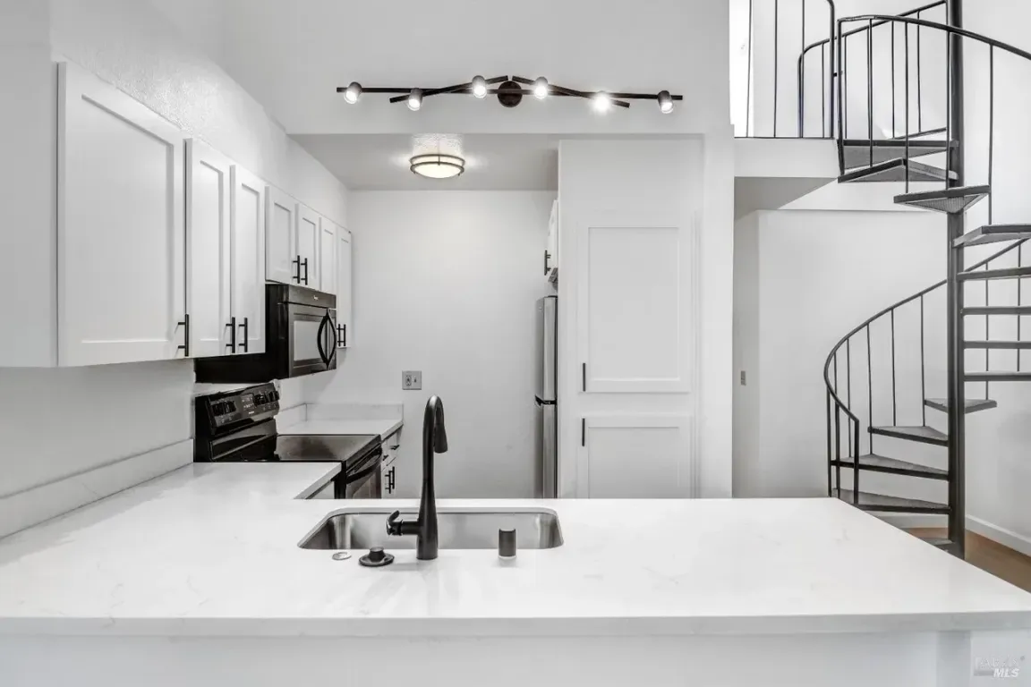 Interior of a Windsor, CA condo kitchen with white cabinets, quartz counters, black fixtures, stainless appliances, and a metal spiral staircase.