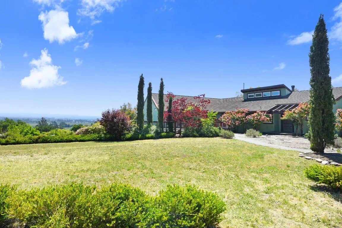 Large country home with tall cypress trees and open lawn under a bright blue sky.