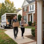 A couple carrying moving boxes toward a moving truck parked in the driveway of a brick suburban home in Charlotte, North Carolina, with a 'Sold' sign in the front yard.