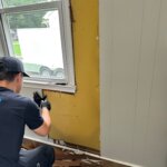 Contractor removing painted wood paneling around a window, revealing the older wall surface and hidden conditions behind it.