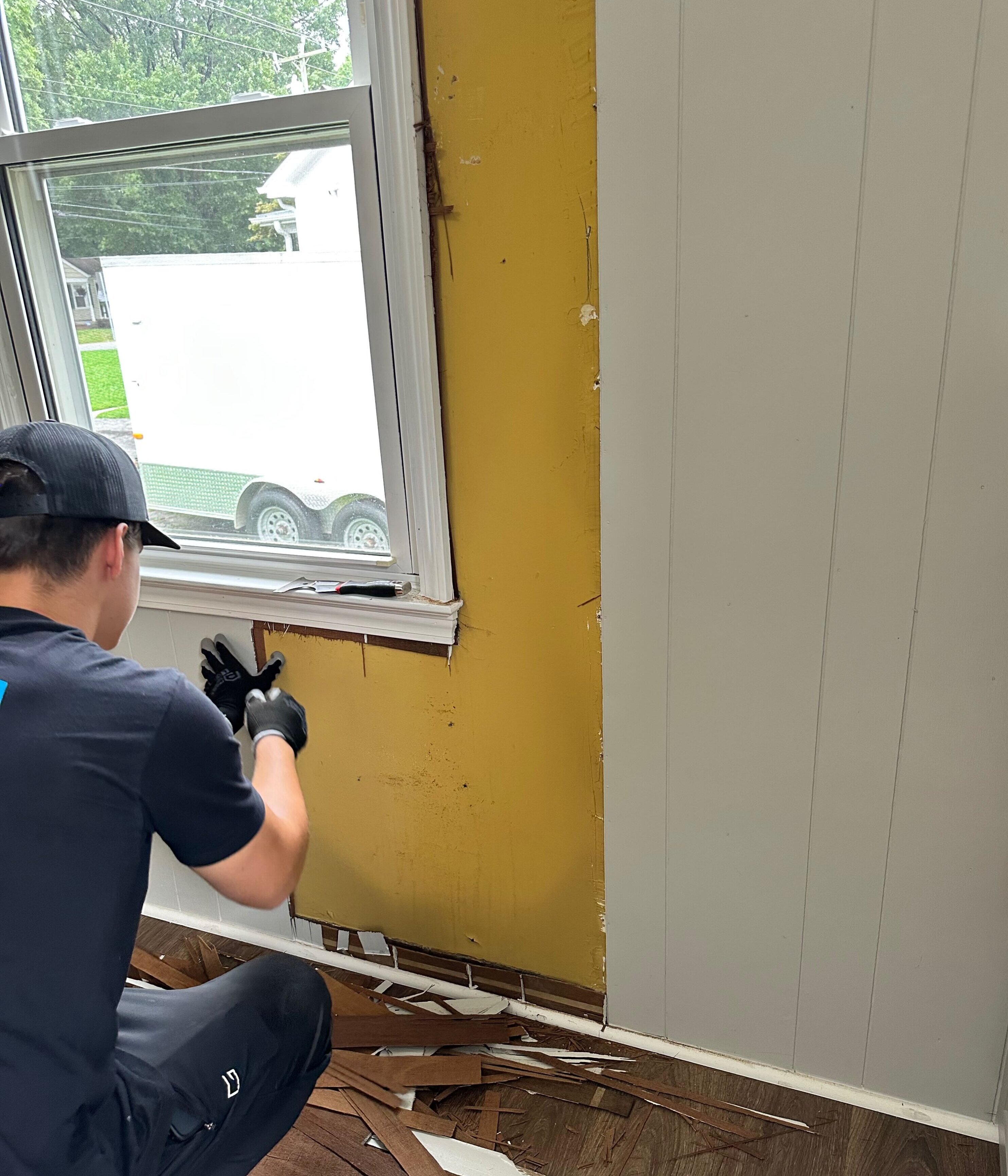 Contractor removing painted wood paneling around a window, revealing the older wall surface and hidden conditions behind it.