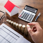 Person Stacking Coins Near House On Wooden Desk Computing Taxes