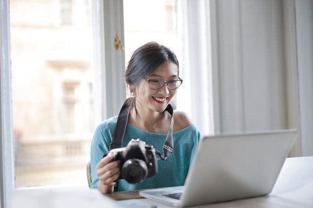 Woman in a green shirt holding a camera while sitting at a desk and looking at a laptop
