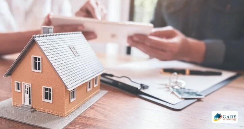 A mini house, documents on the table, and two blurred people in the background.