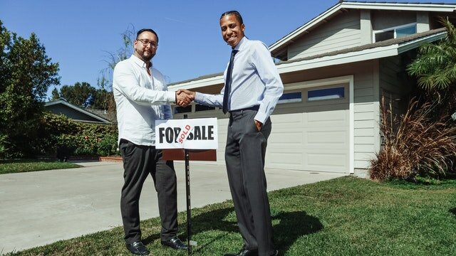 Two people shaking hands in front of a house, next to a sign showing the house has been sold