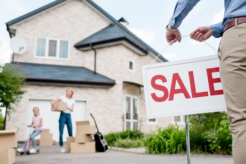 cropped image of male realtor with sale signboard and family with cardboard boxes behind