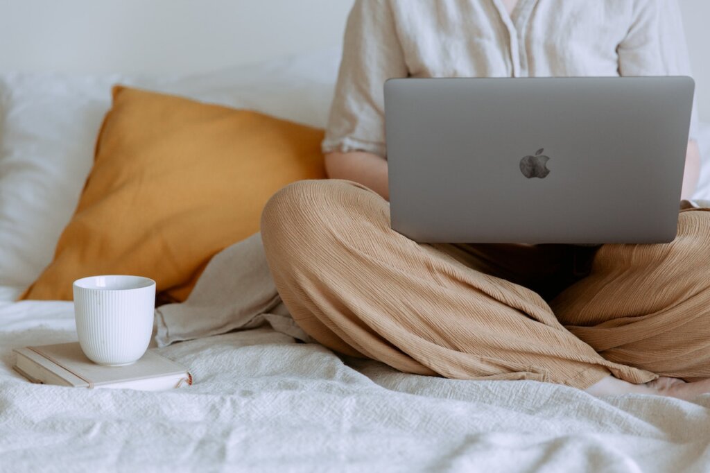 A person sitting on the bed with a laptop in her lap and a cup next to them.