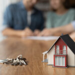 A couple signing house papers in the background, with a scale model and keys representing the real estate transaction in the foreground.