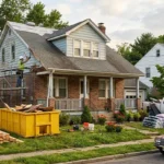 A construction crew renovates a run-down brick and blue-sided house in New Jersey, with scaffolding, a yellow dumpster, and new materials in the yard.