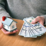local home buyers hands holding new hundred-dollar bills and toy house
