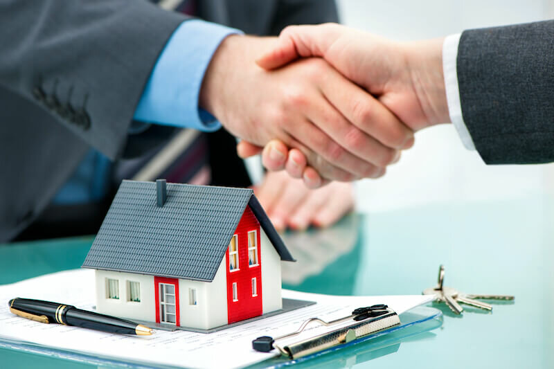 model house and documents with cash home buyer and seller shake hands as background
