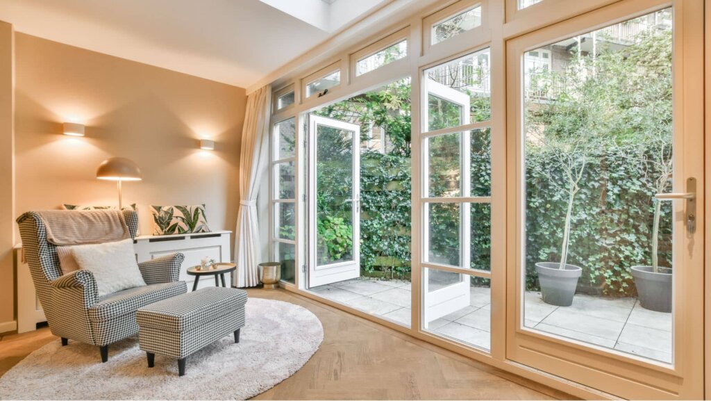 Bright living room with a cozy armchair, ottoman, floor lamp, and side table on a round rug. Large glass doors open to a patio with potted plants—an ideal example of how to enhance property features for homebuyers. Warm lighting creates an inviting atmosphere.