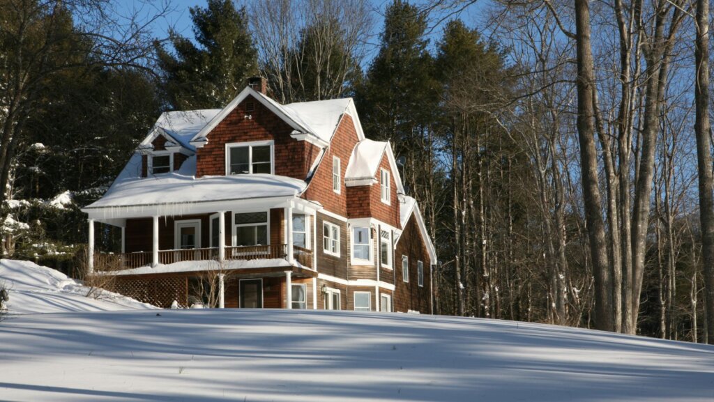 A large, brown wooden house with white trim sits surrounded by snow and tall bare trees, sunlight casting shadows on the snowy ground—an ideal setting if youre considering selling your house for cash this winter.