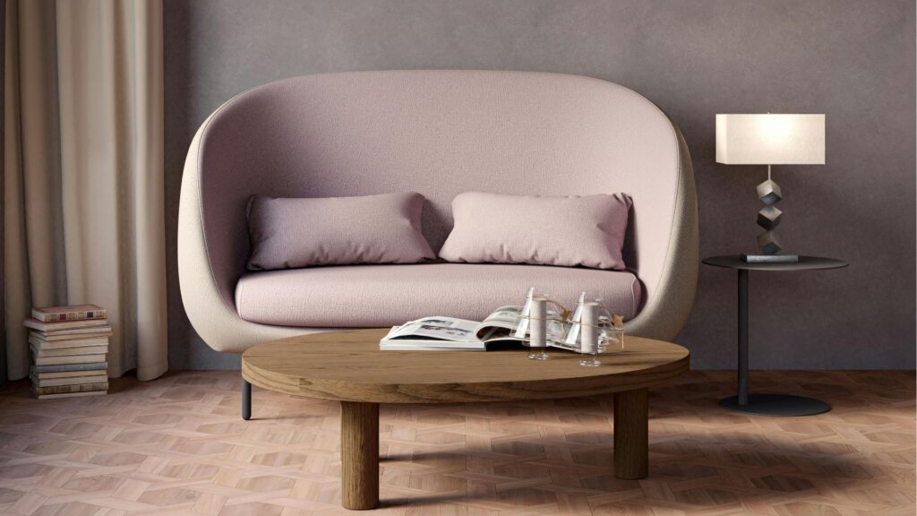 A minimalist living room perfect for a downsizer, featuring a light pink sofa, two cushions, a round wooden coffee table with magazines, a floor lamp, and beige curtains against a gray wall to help you downsize and simplify your life.