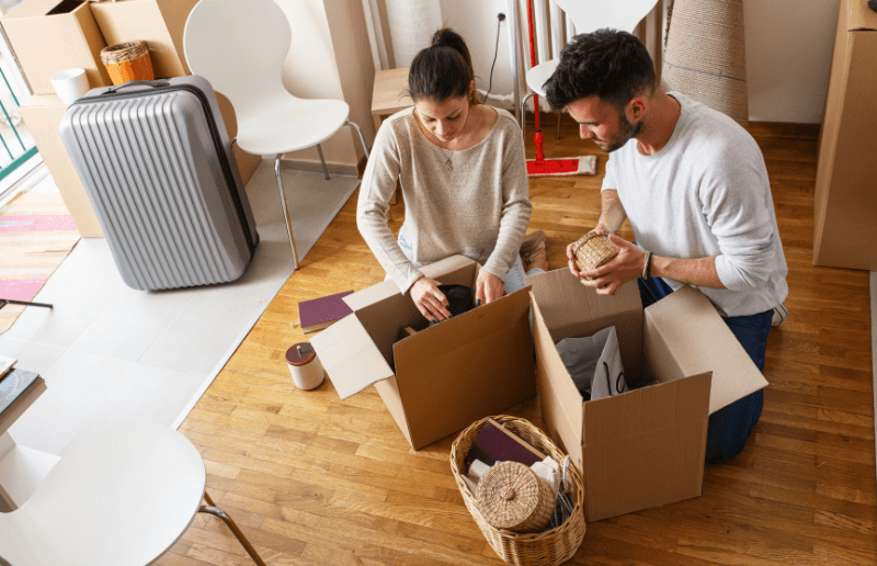 Two young individuals sit down and pack items in boxes while cleaning out a hoarder's house