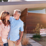 Senior Couple Standing Outdoors In Front Of House With For Sale Sign In Garden