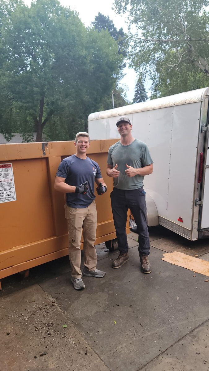 Two people standing beside an orange dumpster and a white trailer.