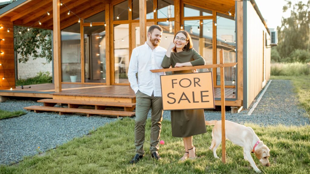 A couple in front of a house holding down a signboard that says "For Sale"