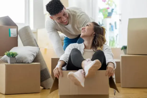 appy couple laughing and playing inside moving boxes in their empty living room after selling their house fast as-is. Cardboard boxes are scattered around, symbolizing a fresh start and stress-free move.