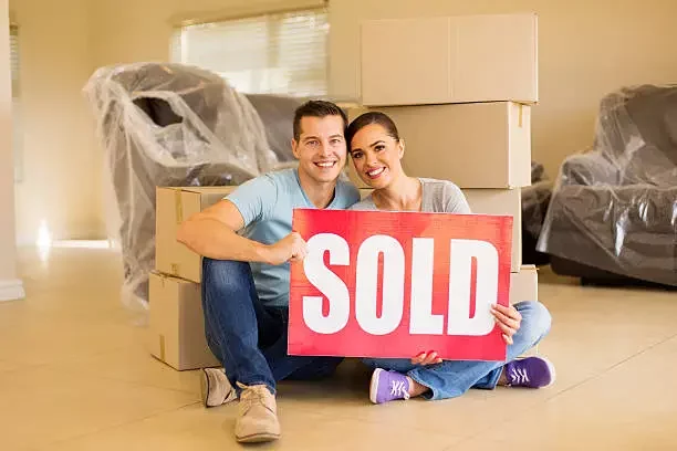 Happy couple sitting on the floor of their recently sold house, smiling and holding a "SOLD" sign, surrounded by moving boxes – celebrating a successful as-is home sale.