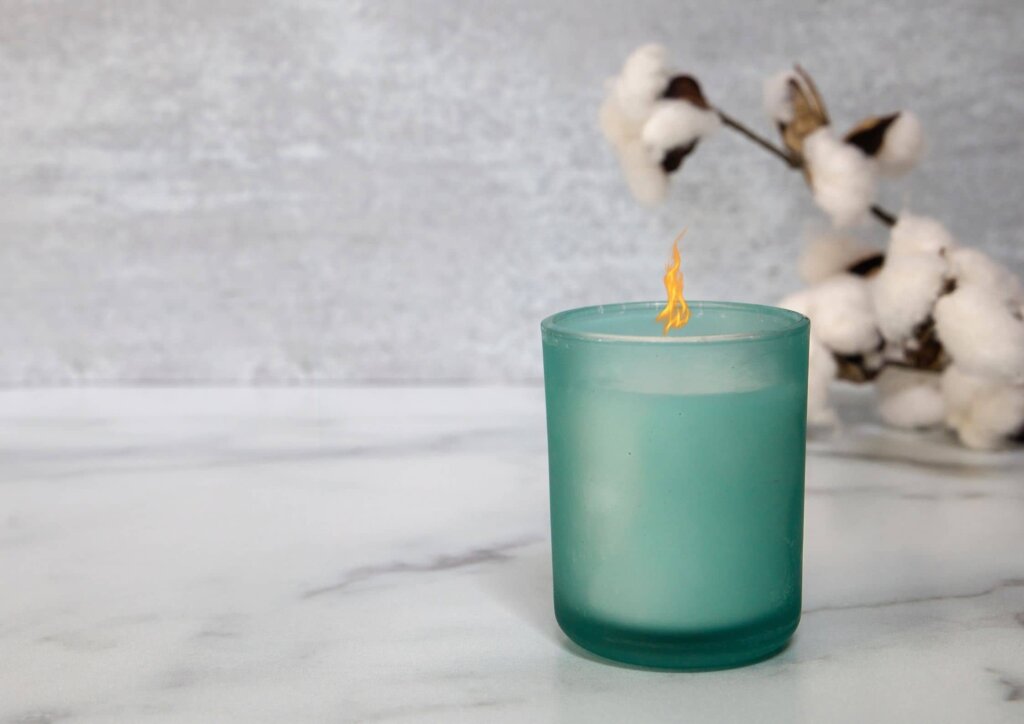 a candle in a blue jar on a marble counter top next to a cotton seed branch