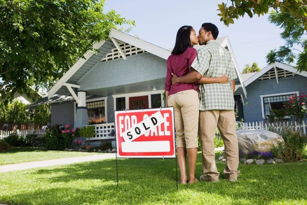 a couple embracing and kissing in front of their house with a sold sign.