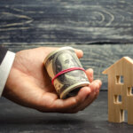 Wooden house and money in hand. A loan secured by real estate. Home renovation and repair. Warm loans. Buying and selling homes. Affordable housing. To rent an apartment. Mortgage rates. Debt. Tax