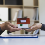 Stock photo for inherited house: two pairs of hands holding a small model house over title transfer paperwork.