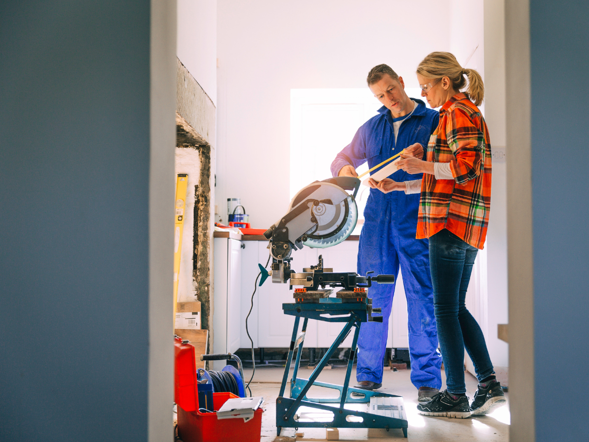 A man and a woman standing in front of a circle saw and measuring a beam of wood.