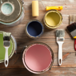 Open paint cans beside paint brushes and paint rollers on a wooden surface.