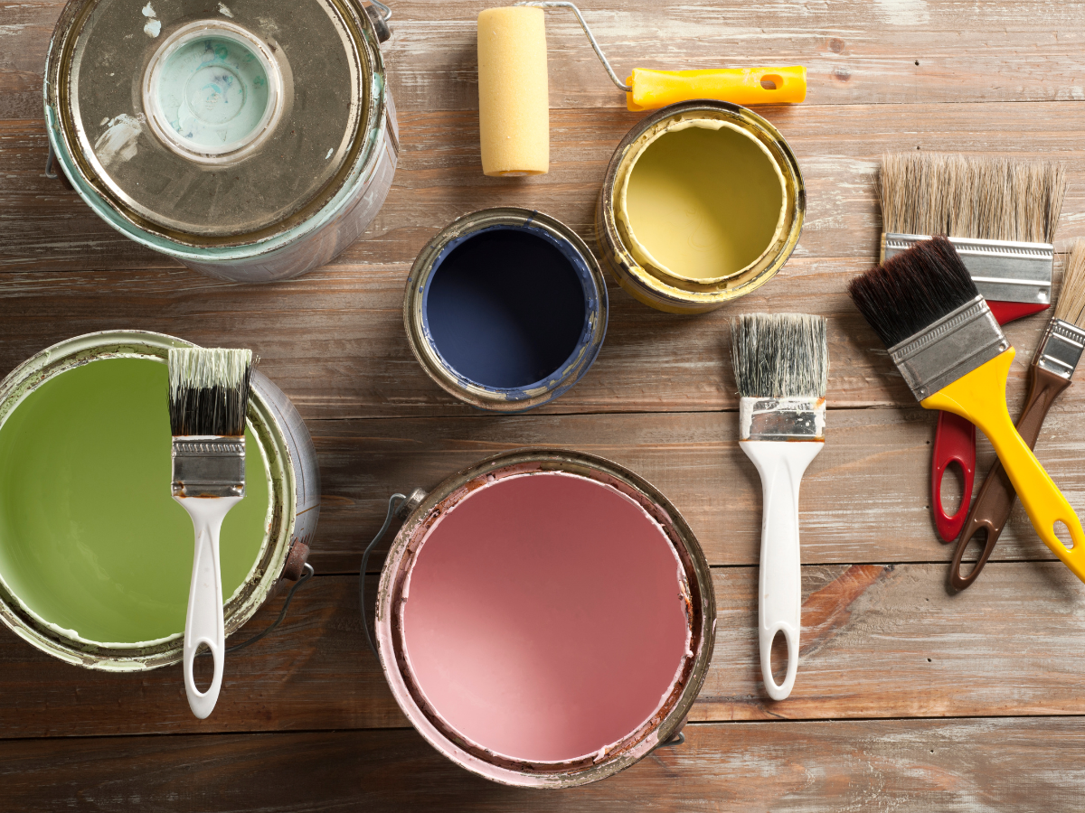 Open paint cans beside paint brushes and paint rollers on a wooden surface.