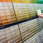 Hands comparing two light paint swatches in front of a wall of multi-colored paint cards in a store.