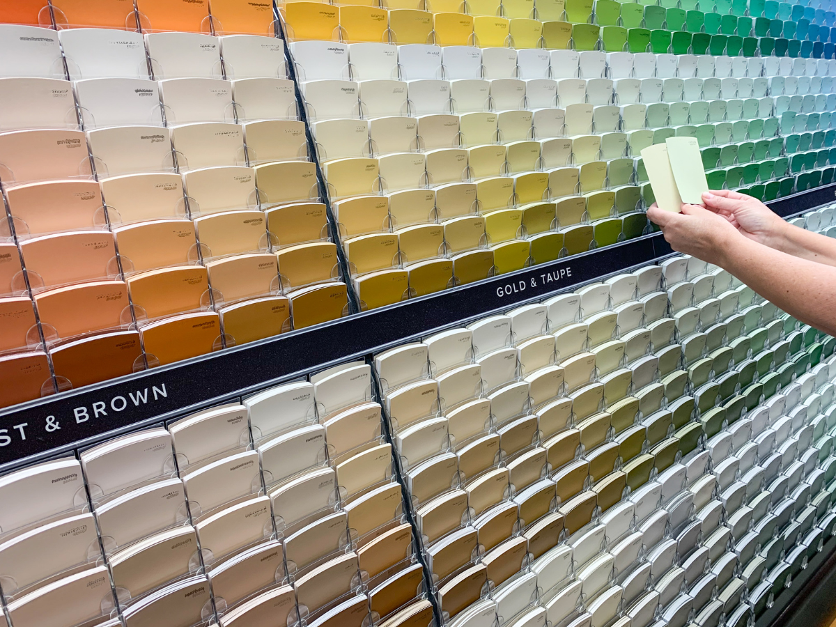 Hands comparing two light paint swatches in front of a wall of multi-colored paint cards in a store.