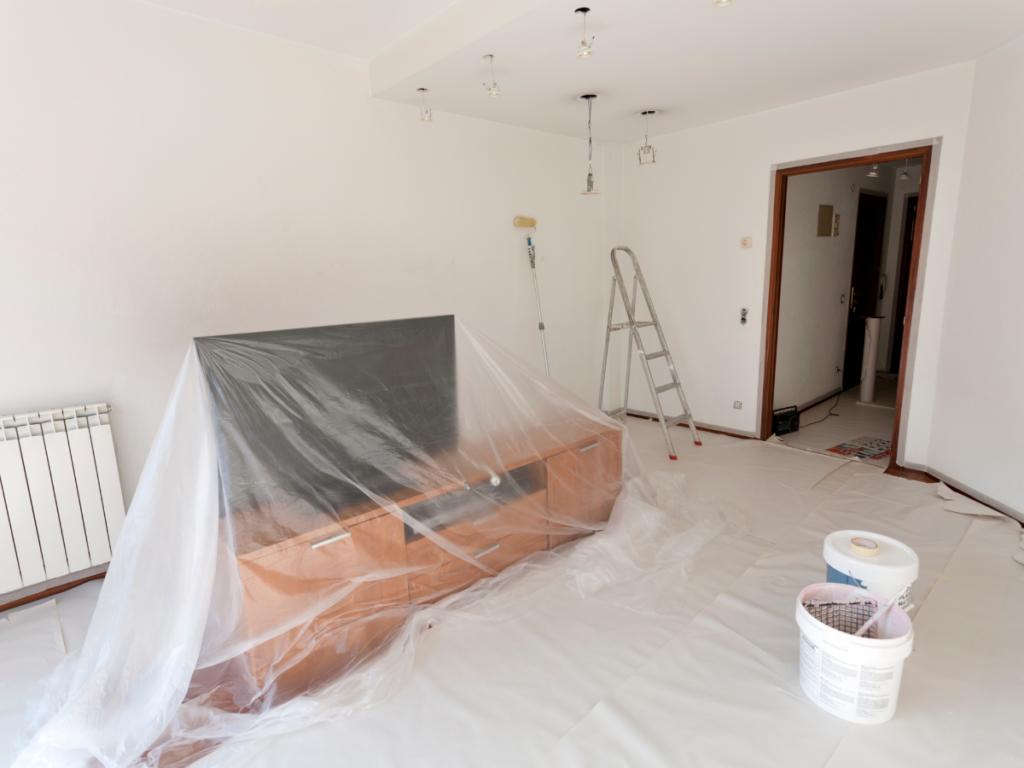 A white room with painting supplies: a roller, a ladder, and 5-gallon buckets. In the center of the room is a TV and TV stand covered with plastic drop cloths.