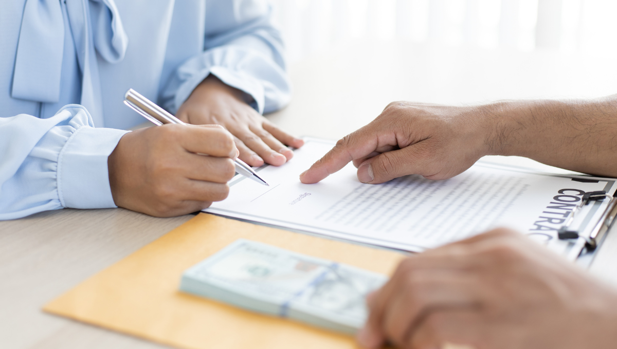 Hands signing a contract beside a stack of money