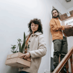 A man and a woman moving packed boxes down a flight of stairs.