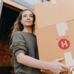 A woman carrying a stack of boxes out of a house.